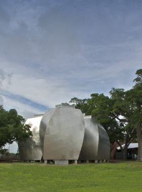 This image shows the modern and artistic design of the Ohr-O’Keefe Museum of Art, featuring futuristic silver buildings designed by Frank Gehry. The unique structures reflect the sunlight, creating a striking visual effect. Visitors are seen walking around the museum, admiring the outdoor sculptures and beautifully landscaped surroundings. Inside, the image shows a display of intricate ceramic pottery pieces, each with bold colors and creative shapes, showcasing the talent of the "Mad Potter of Biloxi," George Ohr.