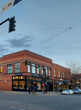 This image shows the vibrant streets of Downtown Bozeman, lined with historic brick buildings, charming cafes, and boutique stores. People can be seen strolling along the sidewalks, enjoying the lively atmosphere. A bookstore with a rustic wooden sign, a cozy coffee shop with outdoor seating, and a local brewery with a welcoming entrance add to the charm. The street is decorated with hanging flower baskets, and a street musician is playing in the background, creating a warm and inviting scene.
