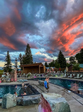 This image shows Bozeman Hot Springs at night, where people are soaking in warm, steamy pools. The outdoor area is illuminated by soft lights, creating a cozy and peaceful atmosphere. The pools vary in size and temperature, with some people enjoying quiet relaxation while others chat with friends. A live band is playing in the background, and the surrounding tall pine trees give the setting a natural, secluded feel, making it the perfect place to unwind.