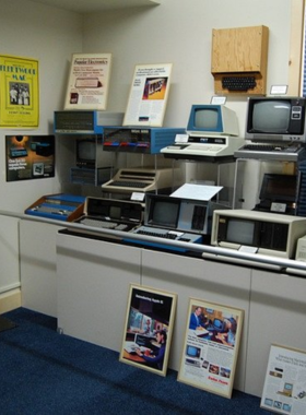 : This image shows an exhibit inside the American Computer & Robotics Museum. Visitors are looking at vintage computers, early calculators, and futuristic robotics displays. An interactive screen provides details about artificial intelligence, while a life-sized robot stands in the corner. The museum has a modern and well-lit interior, with historical artifacts arranged neatly behind glass cases.