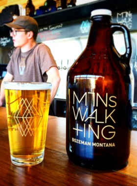 This image shows the cozy interior of Mountains Walking Brewery, where people are enjoying craft beers. A bartender is pouring a golden IPA into a glass, while customers chat at the rustic wooden bar. Large brewing tanks are visible in the background, adding an authentic touch. The ambiance is warm and inviting, with soft lighting and a mix of indoor and outdoor seating.