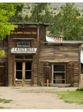 This image shows a group of people on a nighttime ghost tour in Bozeman. A guide holding a lantern is narrating a spooky story in front of an old historic building. The streetlights cast long shadows, and the darkened windows of the buildings add to the eerie atmosphere. The group looks intrigued, some holding cameras to capture the moment.