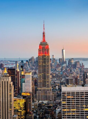 This image shows the breathtaking sunset view from the Empire State Building, one of the most iconic skyscrapers in New York City. The sky is painted in warm hues of orange and pink as the sun sets behind the skyline. Visitors are seen enjoying the view from the observation deck, capturing photos of the city lights beginning to twinkle. The tall skyscrapers, including the Chrysler Building and One World Trade Center, stand in the distance, creating a mesmerizing cityscape. The Empire State Building has been a symbol of NYC since 1931, offering one of the best panoramic views in the city. The deck is crowded with people who have come to witness this magical moment. The glass panels reflect the warm glow of the sunset, making the experience even more spectacular. This image perfectly captures the beauty and grandeur of New York City from one of its most famous landmarks.