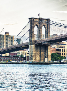 This image shows the Brooklyn Bridge’s pedestrian pathway filled with people walking, jogging, and taking in the stunning views of New York City. The bridge’s towering steel cables and stone arches create a historic and artistic setting. In the background, the Manhattan skyline rises with its towering skyscrapers, while the East River flows beneath the bridge. The wooden plank walkway, built above vehicle lanes, provides a safe and picturesque route for pedestrians and cyclists. The bridge, which was completed in 1883, remains one of the most recognizable landmarks in the city. Some tourists are stopping to take photos, while others are simply enjoying the fresh air and panoramic scenery. The warm golden light of the sunset enhances the beauty of the bridge, creating a peaceful and memorable experience. The Brooklyn Bridge walk is a must-do activity for anyone visiting New York, offering history, exercise, and breathtaking views all in one.