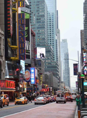 This image shows the vibrant atmosphere of Broadway at night. The street is illuminated with bright, colorful lights from large billboards advertising famous musicals and plays like The Lion King and Hamilton. People are seen walking excitedly towards the theaters, while others are taking pictures of the dazzling surroundings. The grand entrance of a Broadway theater is visible, with its glowing marquee and posters of upcoming shows. The lively crowd and the energy of the place create an unforgettable experience for theater lovers, making Broadway a top destination for entertainment in New York City.