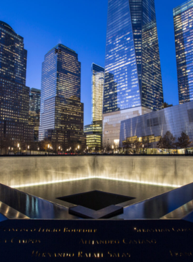 This image shows the 9/11 Memorial reflecting pools, built in the footprints of the original Twin Towers. The cascading waterfalls create a somber and peaceful atmosphere, while engraved names of the victims surround the edges. Visitors stand in silence, paying their respects. In the background, the new One World Trade Center skyscraper rises, symbolizing resilience and hope for the future.