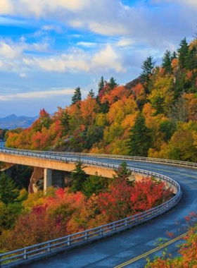 This image shows a breathtaking view of the Blue Ridge Parkway, a winding scenic road stretching through the mountains, covered in dense forests with vibrant fall foliage. The curvy road leads travelers through picturesque landscapes, offering panoramic views of rolling hills, distant peaks, and clear blue skies. This historic parkway provides a peaceful escape into nature, ideal for road trips and photography.