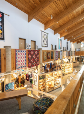 This image shows a beautifully arranged exhibit inside the Folk Art Center, featuring handmade Appalachian crafts such as pottery, quilts, and wooden sculptures. The walls are lined with traditional artwork, while artisans demonstrate their skills in a live crafting session. Visitors walk through the gallery, appreciating the cultural heritage and craftsmanship of the region.