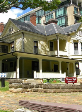 This image shows the historic Thomas Wolfe Memorial House, a well-preserved 19th-century home with a wooden exterior and a wraparound porch. The house, once the childhood home of the famous writer, is surrounded by lush green trees. Visitors can be seen entering the house, eager to explore the literary history and personal artifacts of Thomas Wolfe.