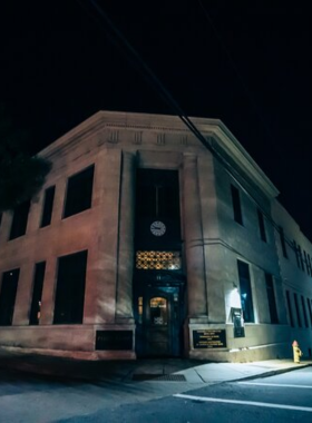 This image shows a nighttime ghost tour in Asheville, where a group of visitors follows a guide dressed in historic attire. The dimly lit streets are lined with old buildings, casting eerie shadows under the streetlights. The tour group listens intently as the guide shares spooky tales of haunted hotels and mysterious events that took place in the city’s past.