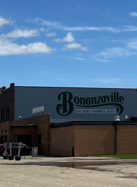 This image shows Bonanzaville, USA, a living history museum in Fargo with authentic 19th-century buildings, including an old general store, a blacksmith shop, and a schoolhouse. Visitors are walking through the dirt paths, stepping inside historic homes, and exploring museum exhibits filled with artifacts from the pioneer days. The scene captures the small-town charm of the Old West, offering a glimpse into life in early North Dakota.