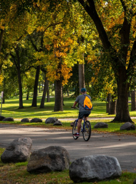 This image shows Lindenwood Park, Fargo’s largest green space, featuring scenic walking trails, a beautiful river, and tall trees providing shade. Families, joggers, and cyclists are enjoying the peaceful outdoor atmosphere, while some visitors are having picnics at the wooden benches and open grassy areas. The park is perfect for relaxation, nature walks, and photography, especially during fall when the trees turn golden and orange.