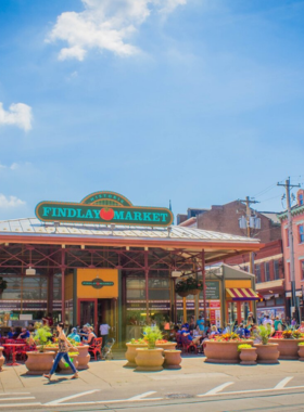 This image shows Findlay Market, Ohio’s oldest continuously operated public market, bustling with vendors and visitors. The vibrant stalls are filled with fresh fruits, vegetables, meats, cheeses, and international street food. Shoppers can be seen enjoying artisanal chocolates, handcrafted goods, and gourmet dishes from local vendors. The lively atmosphere includes street musicians playing cheerful tunes, friendly vendors engaging with customers, and colorful market decor. The market is a cultural hotspot in Cincinnati, where locals and tourists gather to experience diverse flavors, buy unique souvenirs, and soak in the energetic environment.