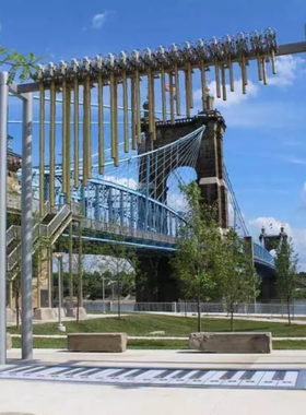 This image shows Smale Riverfront Park, a beautiful urban park along the Ohio River, offering a peaceful retreat in downtown Cincinnati. The park features lush green lawns, scenic walking trails, and people relaxing on giant swings facing the river. Visitors are seen enjoying picnics, children playing in the interactive fountains, and couples taking a stroll along the riverbank. The iconic Roebling Suspension Bridge serves as a stunning backdrop, enhancing the park’s beauty. The setting sun casts a golden glow over the park, creating a magical atmosphere perfect for unwinding, sightseeing, or enjoying outdoor activities.