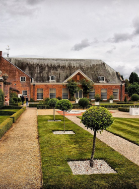 This image shows the grand red-brick facade of Tredegar House, a beautifully preserved 17th-century mansion in Newport, UK. The historic building is surrounded by 90 acres of lush gardens, adding to its charm. The entrance features large wooden doors and classic windows, showcasing its architectural elegance. The house has a rich history, having served as a family home, school, and boarding house, and is now open to visitors who explore its luxurious rooms and gardens.