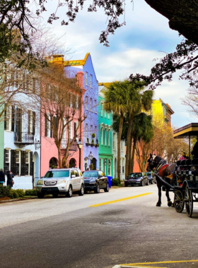 This image shows the historic streets of downtown Charleston, featuring cobblestone roads and colorful 18th-century buildings. The area is rich in history, showcasing charming homes, boutiques, and iconic landmarks. The streets are lined with vibrant flowers and greenery, offering visitors a scenic walk through the heart of Charleston. People enjoy the charming atmosphere as they explore Rainbow Row, a collection of beautifully restored historic homes, and King Street’s shopping district.