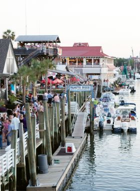 This image shows Shem Creek in Charleston, a lively waterfront area known for its seafood restaurants and shrimp boats. The image features a restaurant patio with diners enjoying fresh seafood dishes while overlooking the creek. Shrimp boats are docked nearby, and visitors can often spot dolphins and other marine life. The picturesque setting provides both delicious meals and scenic views, making it a popular spot for locals and tourists alike to enjoy the coastal charm of Charleston.