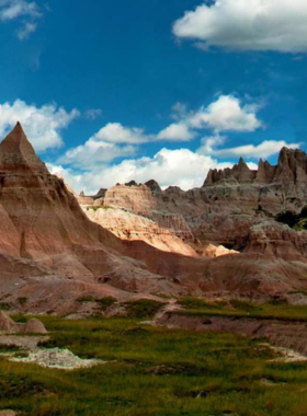 This image shows the breathtaking landscape of Badlands National Park in South Dakota, featuring layered rock formations, dramatic canyons, and sprawling prairies under a clear blue sky. The rugged cliffs display unique shades of red, orange, and brown, shaped by millions of years of erosion. Wildlife such as bison and prairie dogs roam freely, adding to the park’s natural beauty. The vast open land and winding trails make it an ideal destination for photographers, hikers, and nature lovers seeking an adventure in a stunning geological wonderland.