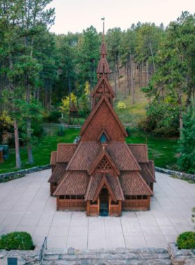 This image shows the beautifully crafted Chapel in the Hills, a wooden church surrounded by lush green trees and a peaceful garden. The chapel’s intricate Scandinavian-style architecture is inspired by Norway’s Borgund Stave Church, featuring wooden carvings, a steep roof, and a historic charm. A small group of visitors stands near the entrance, admiring the peaceful atmosphere. Sunlight filters through the trees, casting a warm glow on the chapel’s exterior, making it a serene and picturesque place for reflection and exploration.