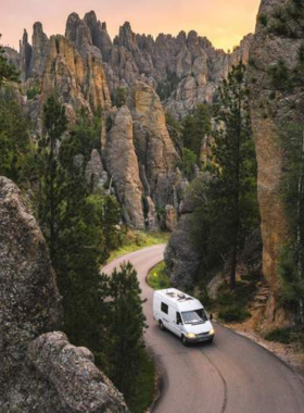 This image shows the winding Needles Highway in South Dakota, cutting through towering granite spires and narrow rock tunnels. A scenic viewpoint overlooks the breathtaking Black Hills landscape, with a clear blue sky and scattered pine trees in the background. A car is driving along the curvy road, adding a sense of adventure and exploration. The rugged rock formations make this a unique and picturesque route, perfect for a memorable road trip with stunning natural views.