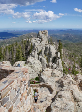 This image shows the breathtaking view from the top of Black Elk Peak, the highest point in South Dakota. A group of hikers stands near the historic fire lookout tower, admiring the panoramic scenery of the Black Hills. The rocky terrain, lush green forests, and rolling hills stretch into the horizon, offering a stunning contrast of colors. Some hikers are seen resting on the rocks, enjoying a well-earned break, while others are taking photos of the incredible landscape. The scene captures the sense of adventure and reward that comes with completing this challenging but scenic trail.