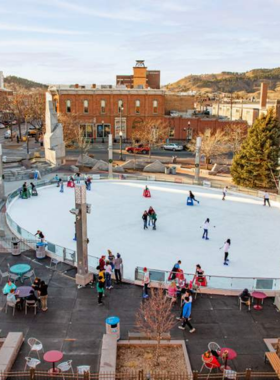 This image shows the lively atmosphere of Main Street Square in Rapid City, with families and children playing in the interactive fountains on a sunny day. The square is surrounded by charming brick buildings with cafes and restaurants, where people can be seen enjoying outdoor dining. In the background, a stage is set up for a live concert, and a crowd gathers to watch the performance. Colorful banners and twinkling lights give the area a festive vibe, making it a vibrant gathering place for locals and tourists alike.