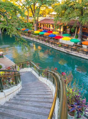 This image shows the stunning San Antonio River Walk at night, illuminated by vibrant lights reflecting on the water. People are seen strolling along the scenic pathways lined with restaurants, bars, and cafes. A boat cruise moves along the river, offering a relaxing ride through the lively area. The atmosphere is warm and inviting, with tourists and locals enjoying delicious food, live music, and a truly unforgettable experience.

