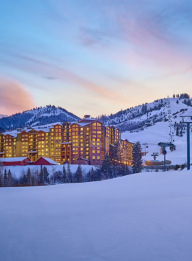 This image shows a breathtaking view of Park City Mountain Resort covered in fresh snow, with skiers and snowboarders gliding down the slopes. The towering mountains in the background are blanketed in white, creating a stunning winter landscape. A ski lift can be seen carrying people up the hill, while others take a break at a cozy lodge. The bright blue sky contrasts with the snowy peaks, making the scene look picturesque. The resort is the largest in the U.S., offering ski terrain for all skill levels. Some skiers are seen practicing on gentle slopes, while experts race down challenging trails. The image captures the joy and excitement of winter sports, highlighting why Park City is a paradise for snow lovers. Snow-covered trees add to the scenic beauty, while people in colorful ski gear add vibrancy to the image.