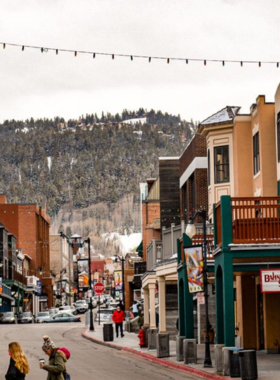 This image shows the charming Historic Main Street in Park City, lined with colorful buildings and twinkling lights. The street has a nostalgic feel with its historic architecture, boutique shops, and local cafes. A couple walks along the sidewalk, sipping coffee and admiring window displays. Art galleries and bookstores can be seen, offering unique finds for visitors. In the evening, the street glows under golden lights, creating a magical atmosphere. Snow-covered rooftops add to the cozy winter vibe. Live music plays from a local bar, drawing a small crowd. The street is filled with people exploring, shopping, and enjoying the lively ambiance. The combination of history, luxury, and small-town charm makes this a must-visit spot. The image perfectly captures the inviting nature of Park City’s Historic Main Street, where old-world charm meets modern entertainment.