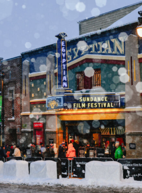 This image shows the bustling atmosphere of the Sundance Film Festival in Park City, with filmmakers, celebrities, and movie enthusiasts gathered for screenings and events. A crowd of excited visitors lines up outside a historic theater, eager to watch an independent film premiere. Snowflakes gently fall, adding to the winter charm of the festival. Bright banners and posters featuring film titles decorate the streets, while photographers capture memorable moments. A filmmaker is seen discussing their work during a Q&A session, engaging with an enthusiastic audience. The event is known for showcasing groundbreaking independent films, and this image highlights its artistic energy. Small cafes and restaurants are filled with people discussing their favorite screenings. The festival brings an electrifying energy to the town, making it a cultural hotspot. The image perfectly captures the excitement and prestige of the Sundance Film Festival, where creativity and storytelling come to life.