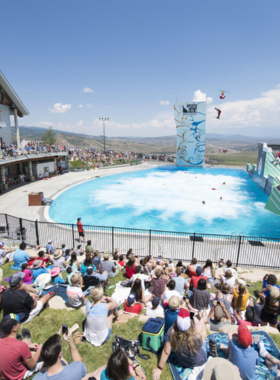 This image showcases Utah Olympic Park, an iconic training and competition venue built for the 2002 Winter Olympics. A massive ski jump tower dominates the scene, with athletes soaring through the air during a practice session. Below, visitors watch in awe, some capturing the moment on their phones. The Comet Bobsled track is also visible, where thrill-seekers experience speeds of up to 70 mph. In the distance, an interactive museum building houses Olympic memorabilia and history exhibits. Adventure seekers enjoy extreme ziplining and ropes courses, adding to the park’s vibrant energy. The park is an ideal place for sports lovers, offering an up-close look at professional training facilities. The image perfectly captures the Olympic spirit, highlighting Park City’s legacy in winter sports and adventure.