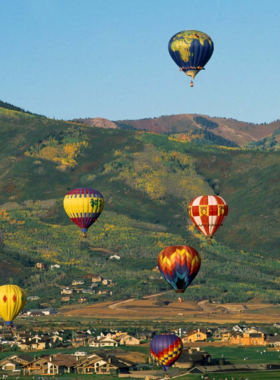 This image showcases a breathtaking hot air balloon ride over Park City, with colorful balloons floating high above the Wasatch Mountains. A bright sunrise paints the sky in shades of pink, orange, and gold, illuminating the rolling hills and valleys below. The perspective from the basket gives a stunning bird’s-eye view of the landscape, with forests, lakes, and snow-capped peaks stretching for miles. A couple stands inside the balloon’s basket, gazing out in awe as they gently rise higher. Another balloon is seen in the distance, adding to the scenic beauty. After landing, a small group of guests celebrates with a traditional champagne toast, marking the end of a magical experience. The image perfectly captures the peaceful yet exhilarating nature of a hot air balloon ride in Park City.