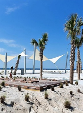 This image shows Gulf Shores Public Beach with its pristine white sand and crystal-clear waters. The tranquil setting captures the peaceful atmosphere of this coastal paradise where families and individuals relax by the ocean. You can spot people lounging under umbrellas, children playing near the gentle waves, and beachside shops and restaurants in the background. The soothing sunset adds to the serene ambiance, making it the perfect spot for relaxation and enjoyment of the Gulf Coast’s beauty.